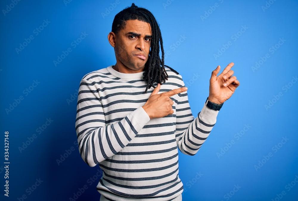 Naklejka premium Young handsome african american afro man with dreadlocks wearing casual striped sweater Pointing aside worried and nervous with both hands, concerned and surprised expression