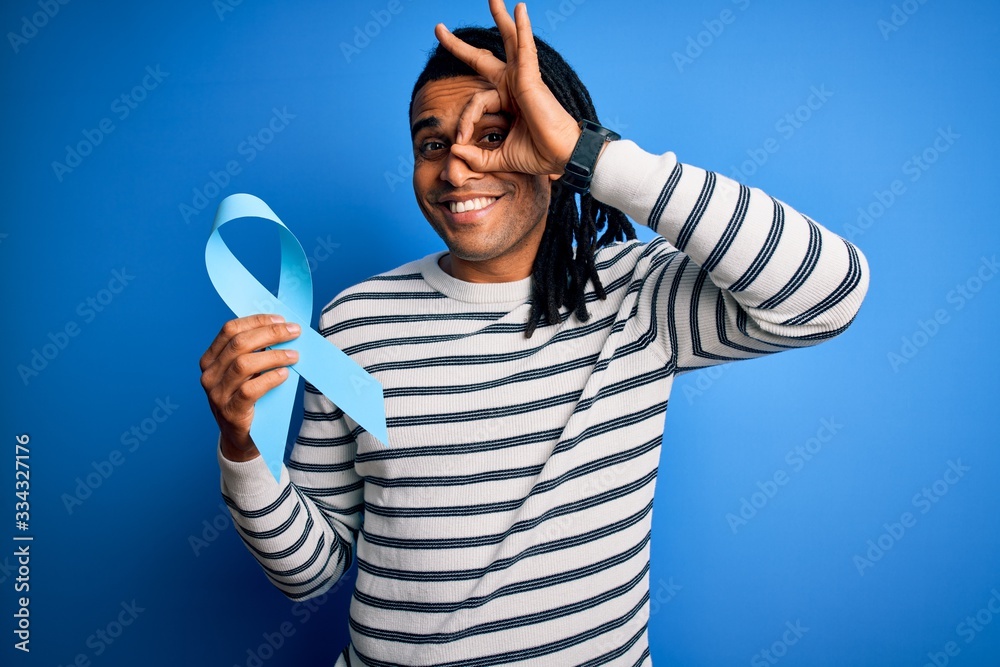 Young african american afro man with dreadlocks holding blue cancer ribbon with happy face smiling doing ok sign with hand on eye looking through fingers Young african american afro man with dreadlocks holding blue cancer ribbon with happy face smiling doing ok sign with hand on eye looking through fingers