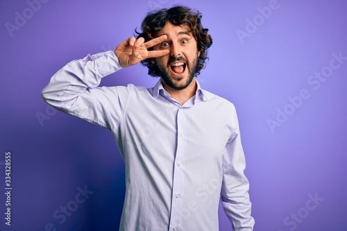 Young handsome business man with beard wearing shirt standing over purple background Doing peace symbol with fingers over face, smiling cheerful showing victory
