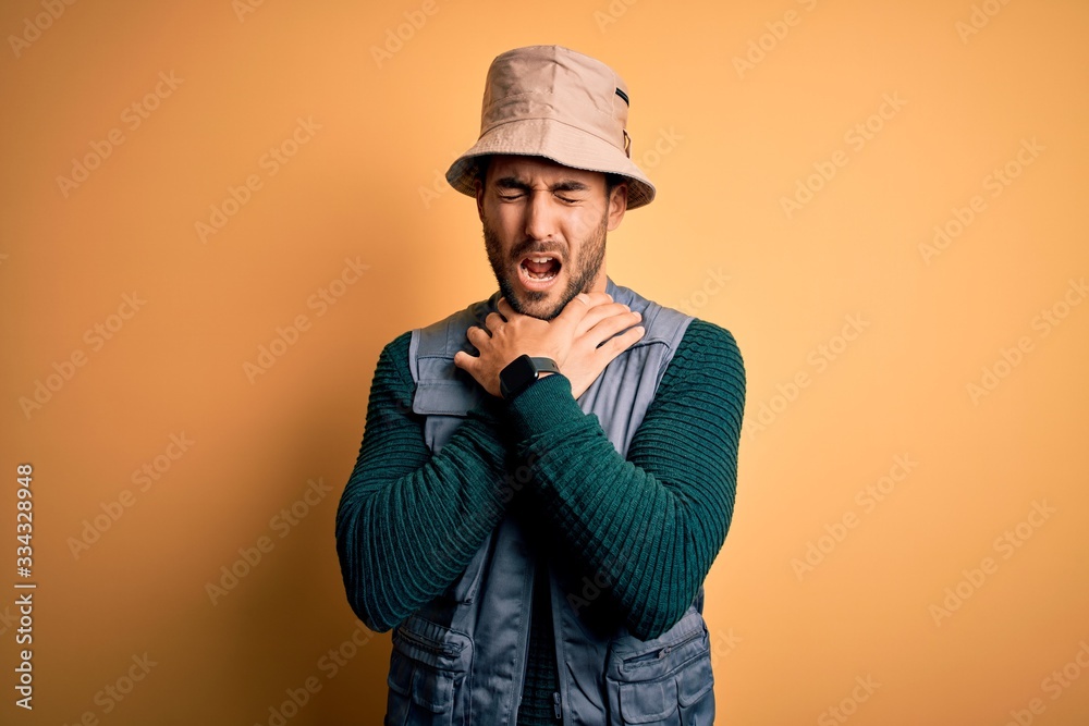 Handsome tourist man with beard on vacation wearing explorer hat over yellow background shouting suffocate because painful strangle. Health problem. Asphyxiate and suicide concept. Handsome tourist man with beard on vacation wearing explorer hat over yellow background shouting suffocate because painful strangle. Health problem. Asphyxiate and suicide concept.