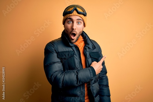Young handsome skier man with beard wearing snow sportswear and ski goggles Surprised pointing with finger to the side, open mouth amazed expression. © Krakenimages.com