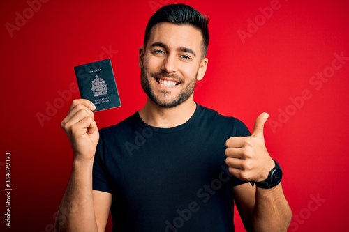 Young handsome tourist man holding canada canadian passport id over red background happy with big smile doing ok sign, thumb up with fingers, excellent sign © Krakenimages.com