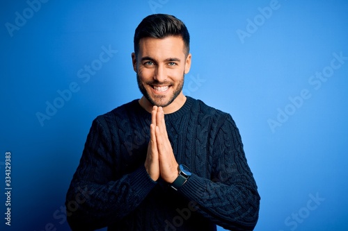 Young handsome man wearing casual sweater standing over isolated blue background praying with hands together asking for forgiveness smiling confident.