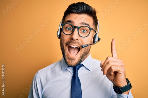 Young business operator man wearing customer service headset from call center pointing finger up with successful idea. Exited and happy. Number one.