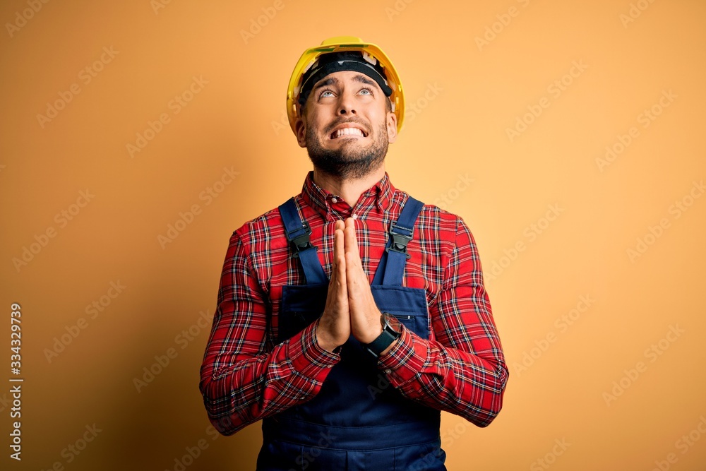 Young builder man wearing construction uniform and safety helmet over yellow isolated background begging and praying with hands together with hope expression on face very emotional and worried Young builder man wearing construction uniform and safety helmet over yellow isolated background begging and praying with hands together with hope expression on face very emotional and worried