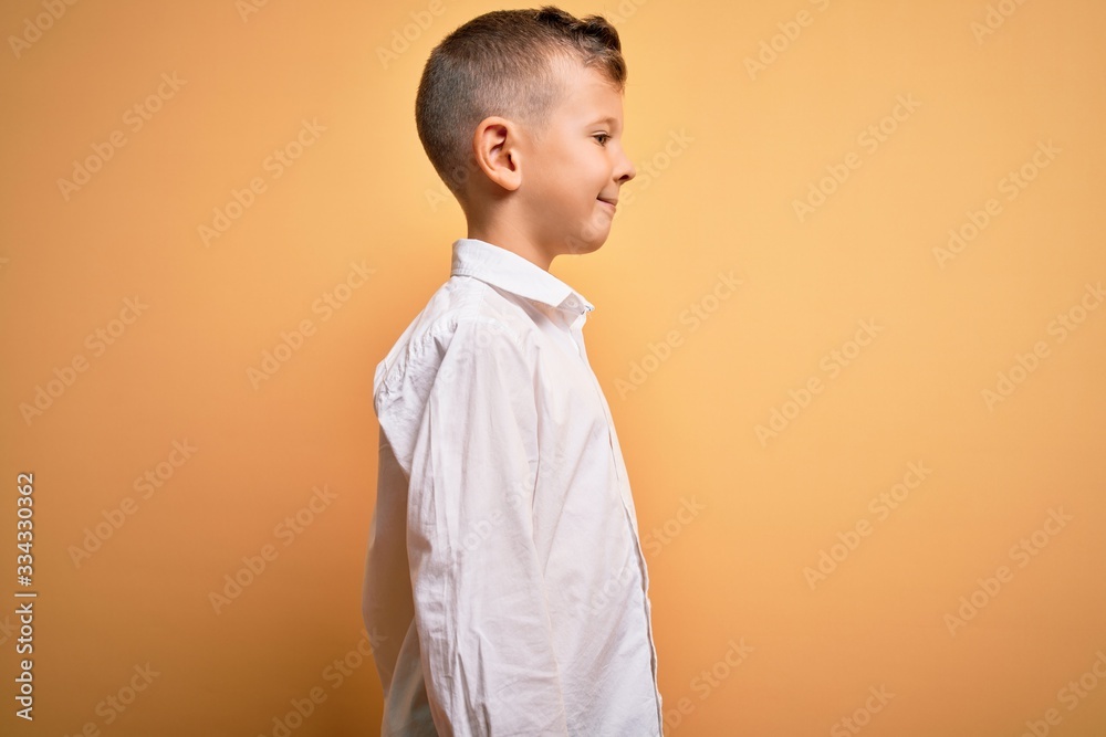 Young little caucasian kid with blue eyes wearing elegant white shirt over yellow background looking to side, relax profile pose with natural face with confident smile. Young little caucasian kid with blue eyes wearing elegant white shirt over yellow background looking to side, relax profile pose with natural face with confident smile.