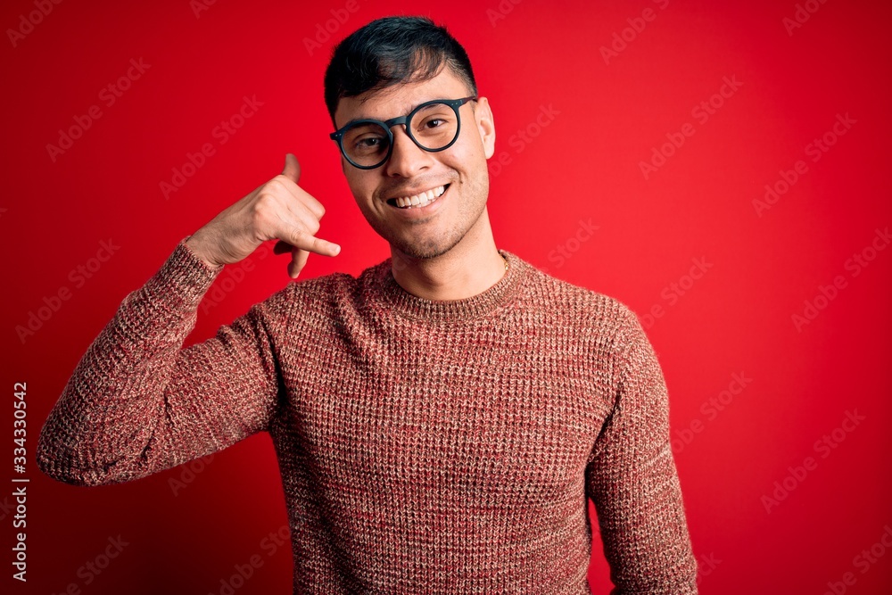Obraz premium Young handsome hispanic man wearing nerd glasses over red background smiling doing phone gesture with hand and fingers like talking on the telephone. Communicating concepts.