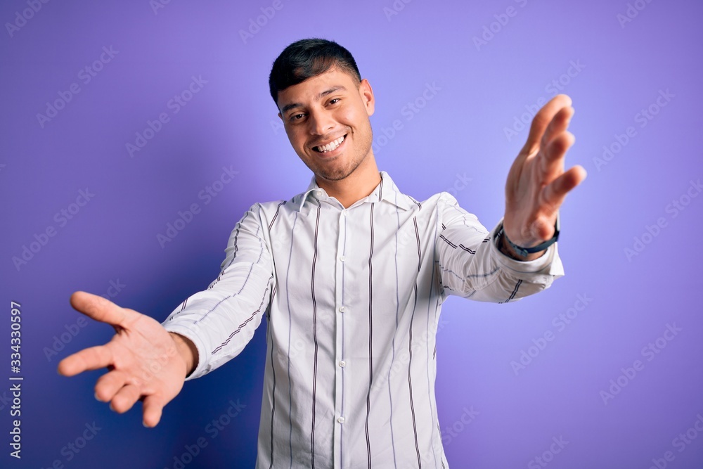 Young handsome hispanic man wearing elegant business shirt standing over purple background looking at the camera smiling with open arms for hug. Cheerful expression embracing happiness. Young handsome hispanic man wearing elegant business shirt standing over purple background looking at the camera smiling with open arms for hug. Cheerful expression embracing happiness.