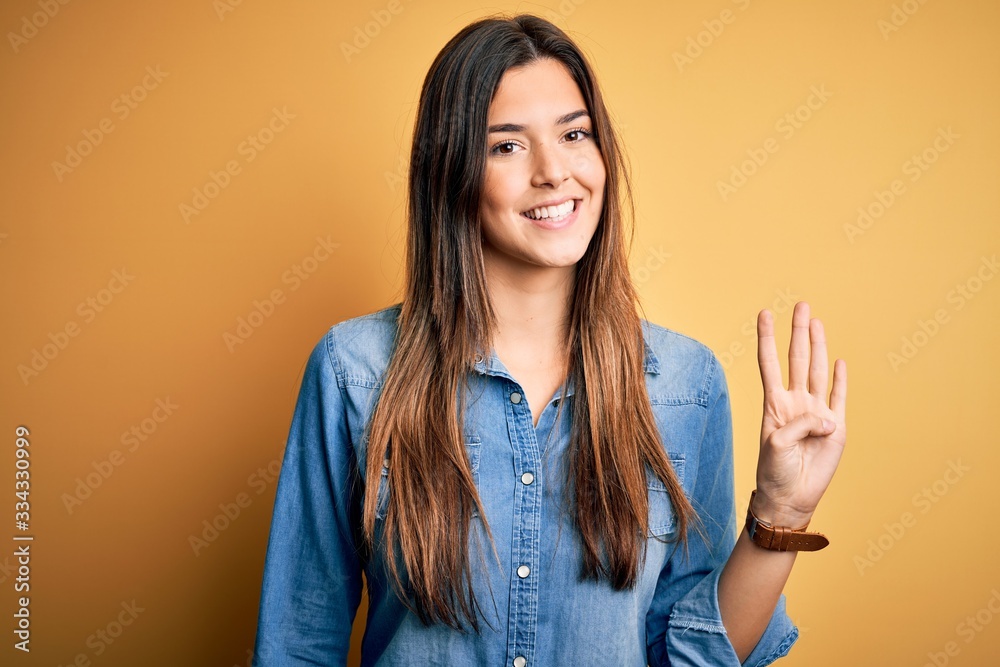 Young beautiful girl wearing casual denim shirt standing over isolated yellow background showing and pointing up with fingers number four while smiling confident and happy. Young beautiful girl wearing casual denim shirt standing over isolated yellow background showing and pointing up with fingers number four while smiling confident and happy.