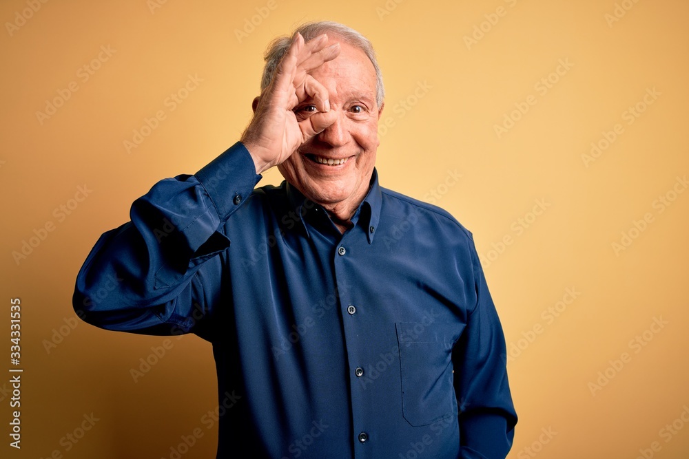 Obraz premium Grey haired senior man wearing casual blue shirt standing over yellow background doing ok gesture with hand smiling, eye looking through fingers with happy face.