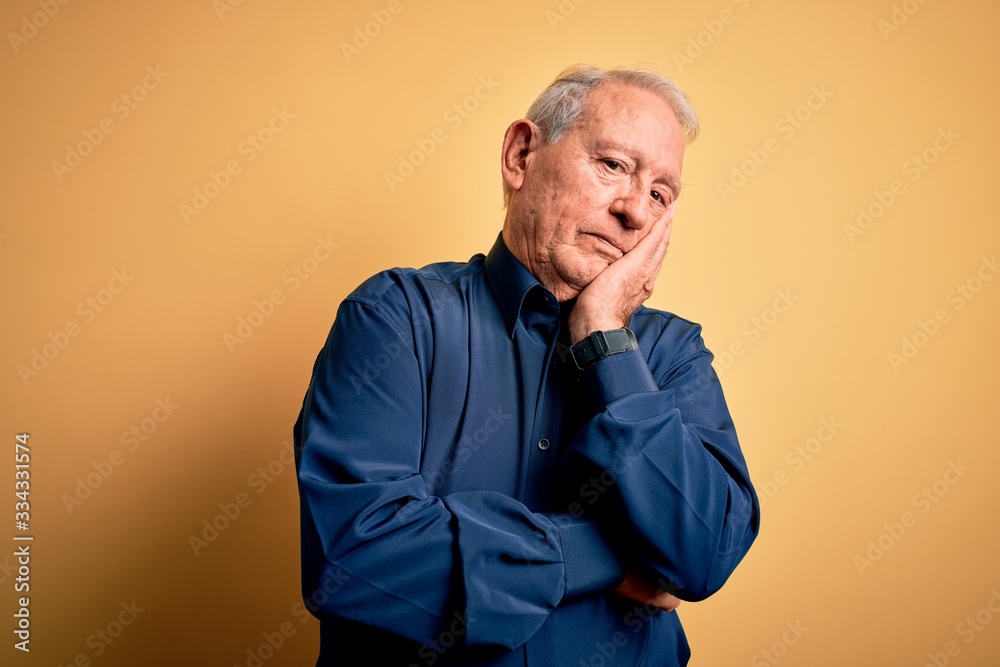 Grey haired senior man wearing casual blue shirt standing over yellow background thinking looking tired and bored with depression problems with crossed arms.