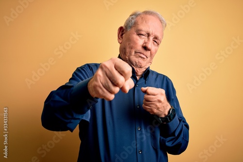 Grey haired senior man wearing casual blue shirt standing over yellow background Punching fist to fight, aggressive and angry attack, threat and violence