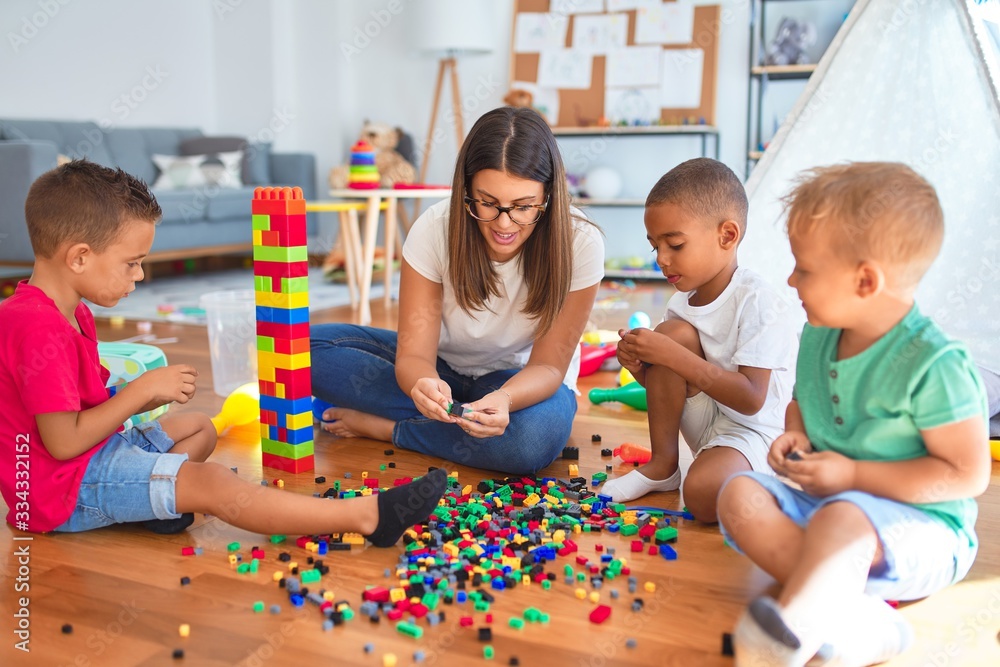 © Krakenimages.com - Young beautiful teacher and toddlers playing with building blocks around lots of toys at kindergarten