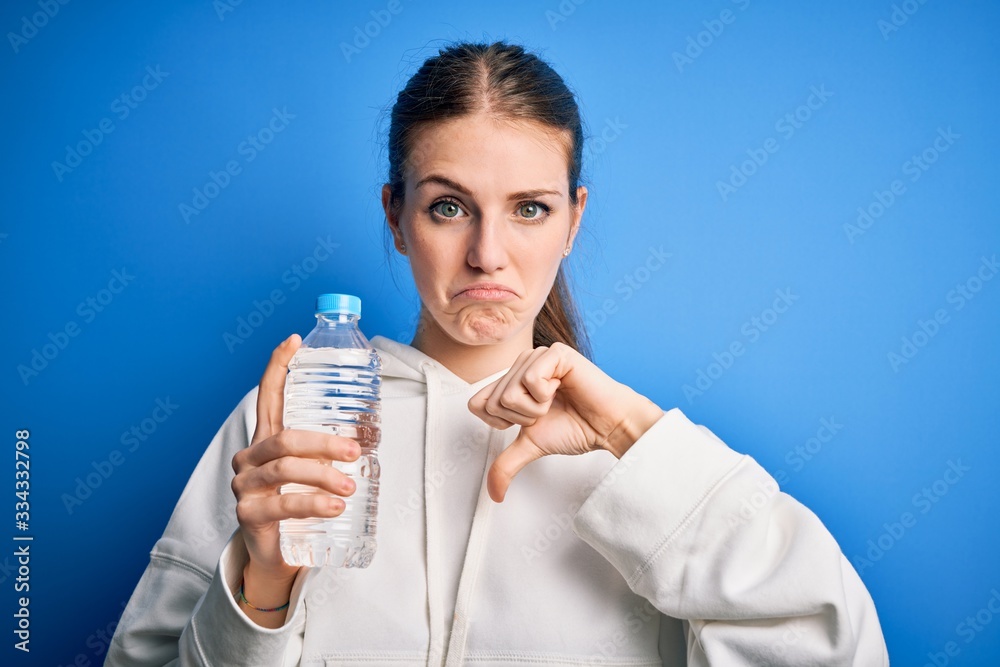 Young beautiful redhead woman doing sport drinking bottle of water over blue background with angry face, negative sign showing dislike with thumbs down, rejection concept Young beautiful redhead woman doing sport drinking bottle of water over blue background with angry face, negative sign showing dislike with thumbs down, rejection concept