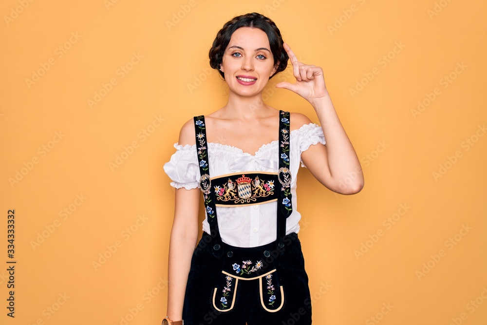 Young beautiful german woman with blue eyes wearing traditional octoberfest dress smiling and confident gesturing with hand doing small size sign with fingers looking and the camera. Measure concept. Young beautiful german woman with blue eyes wearing traditional octoberfest dress smiling and confident gesturing with hand doing small size sign with fingers looking and the camera. Measure concept.