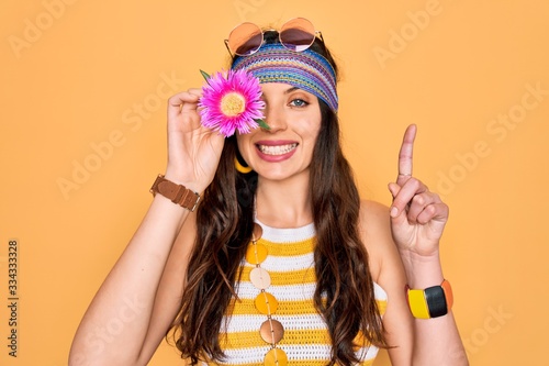 Young beautiful hippie woman with blue eyes wearing sunnglasses holding pink flower over eye surprised with an idea or question pointing finger with happy face, number one
