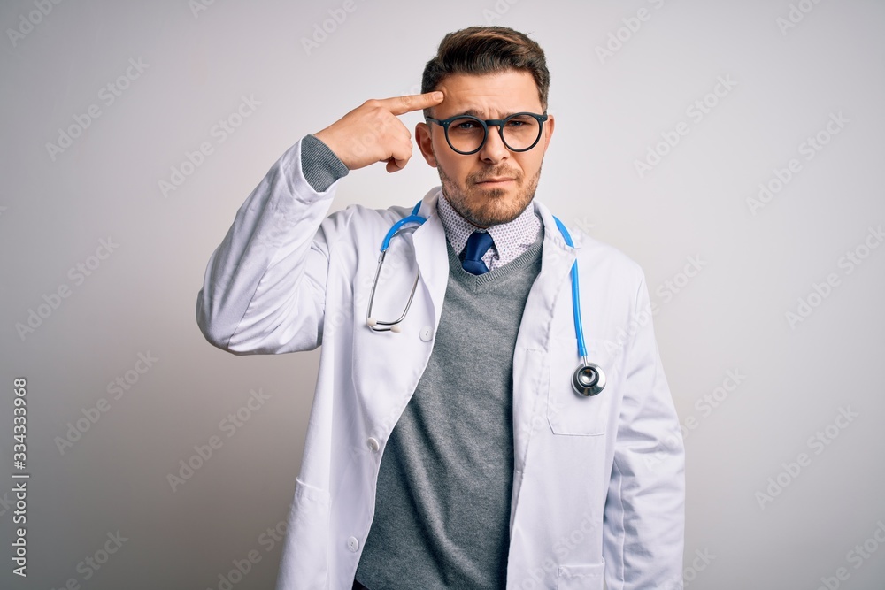 Young doctor man with blue eyes wearing medical coat and stethoscope over isolated background pointing unhappy to pimple on forehead, ugly infection of blackhead. Acne and skin problem Young doctor man with blue eyes wearing medical coat and stethoscope over isolated background pointing unhappy to pimple on forehead, ugly infection of blackhead. Acne and skin problem