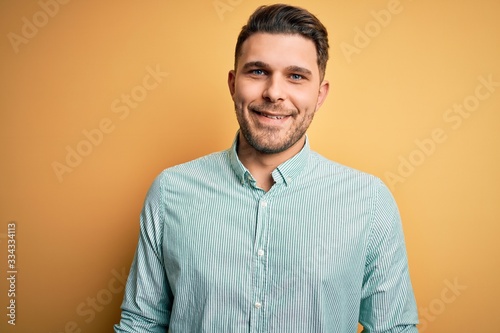 Young business man with blue eyes wearing elegant green shirt over yellow background with a happy and cool smile on face. Lucky person.