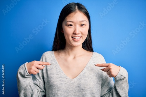 Young beautiful asian woman wearing casual sweater standing over blue isolated background looking confident with smile on face, pointing oneself with fingers proud and happy.