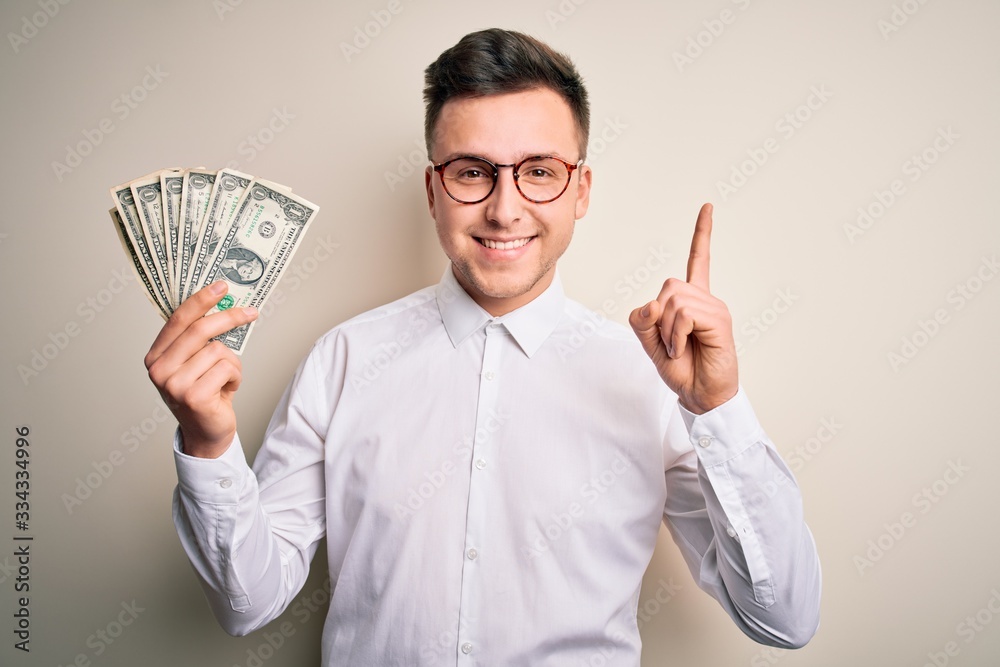 Young handsome caucasian business man holding bunch of one dollar banknotes surprised with an idea or question pointing finger with happy face, number one Young handsome caucasian business man holding bunch of one dollar banknotes surprised with an idea or question pointing finger with happy face, number one