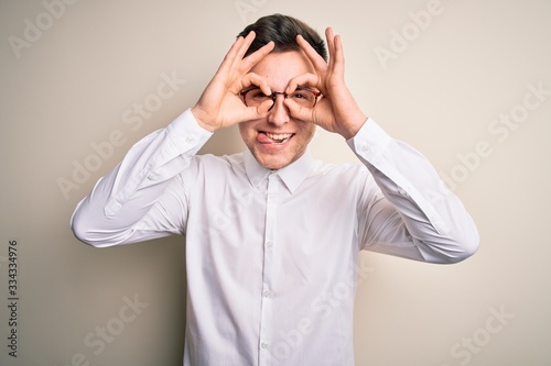 Young handsome business mas wearing glasses and elegant shirt over isolated background doing ok gesture like binoculars sticking tongue out, eyes looking through fingers. Crazy expression.