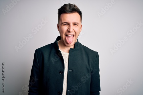 Young handsome business mas wearing elegant winter coat standing over isolated background sticking tongue out happy with funny expression. Emotion concept.