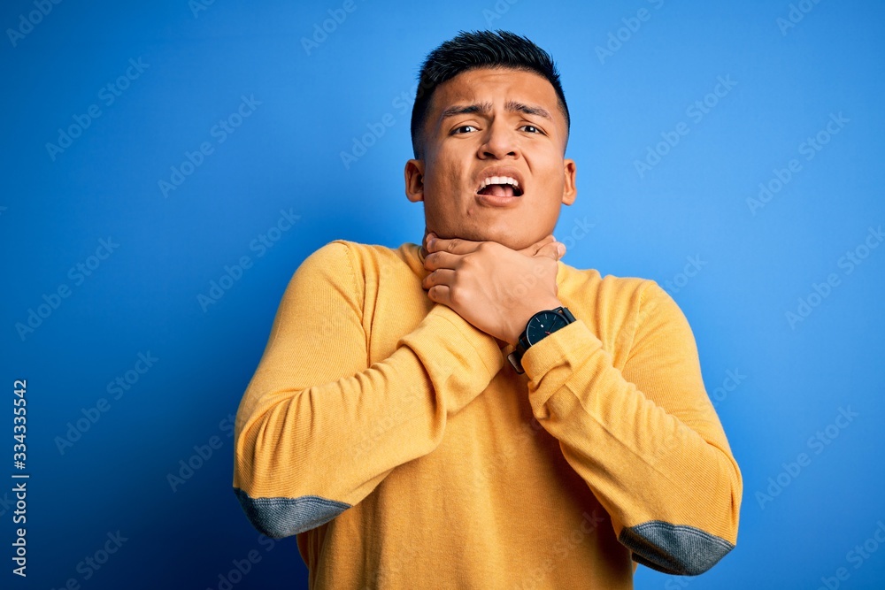 Young handsome latin man wearing yellow casual sweater over isolated blue background shouting suffocate because painful strangle. Health problem. Asphyxiate and suicide concept. Young handsome latin man wearing yellow casual sweater over isolated blue background shouting suffocate because painful strangle. Health problem. Asphyxiate and suicide concept.