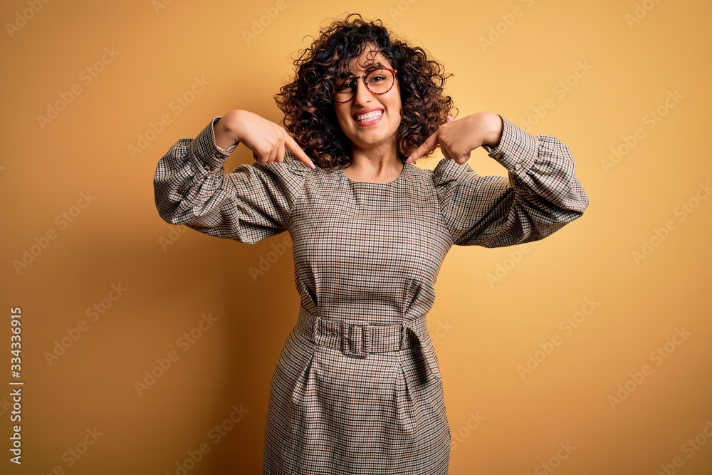 Beautiful arab business woman wearing dress and glasses standing over yellow background looking confident with smile on face, pointing oneself with fingers proud and happy.