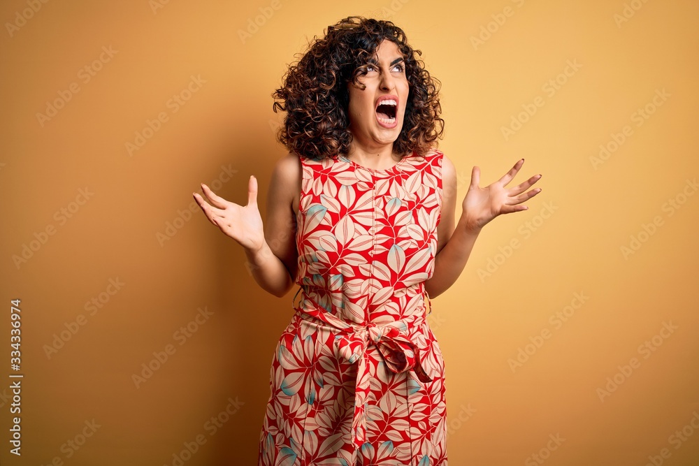 Young beautiful curly arab woman on vacation wearing summer floral dress and sunglasses crazy and mad shouting and yelling with aggressive expression and arms raised. Frustration concept. Young beautiful curly arab woman on vacation wearing summer floral dress and sunglasses crazy and mad shouting and yelling with aggressive expression and arms raised. Frustration concept.