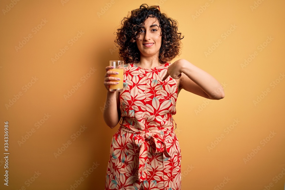 Young beautiful curly arab woman drinking glass of healthy orange juice over yellow background with surprise face pointing finger to himself Young beautiful curly arab woman drinking glass of healthy orange juice over yellow background with surprise face pointing finger to himself