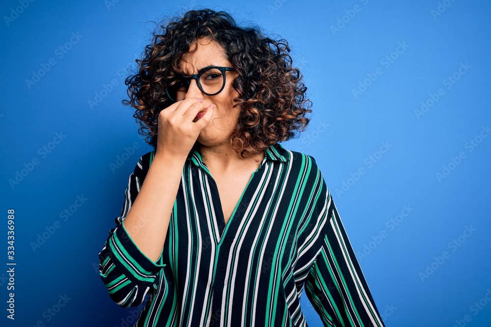 Young beautiful curly arab woman wearing striped shirt and glasses over blue background smelling something stinky and disgusting, intolerable smell, holding breath with fingers on nose. Bad smell