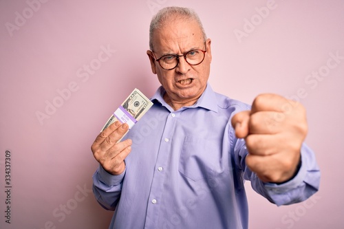 Middle age hoary man holding bunch of dollars banknotes over isolated pink background annoyed and frustrated shouting with anger, crazy and yelling with raised hand, anger concept