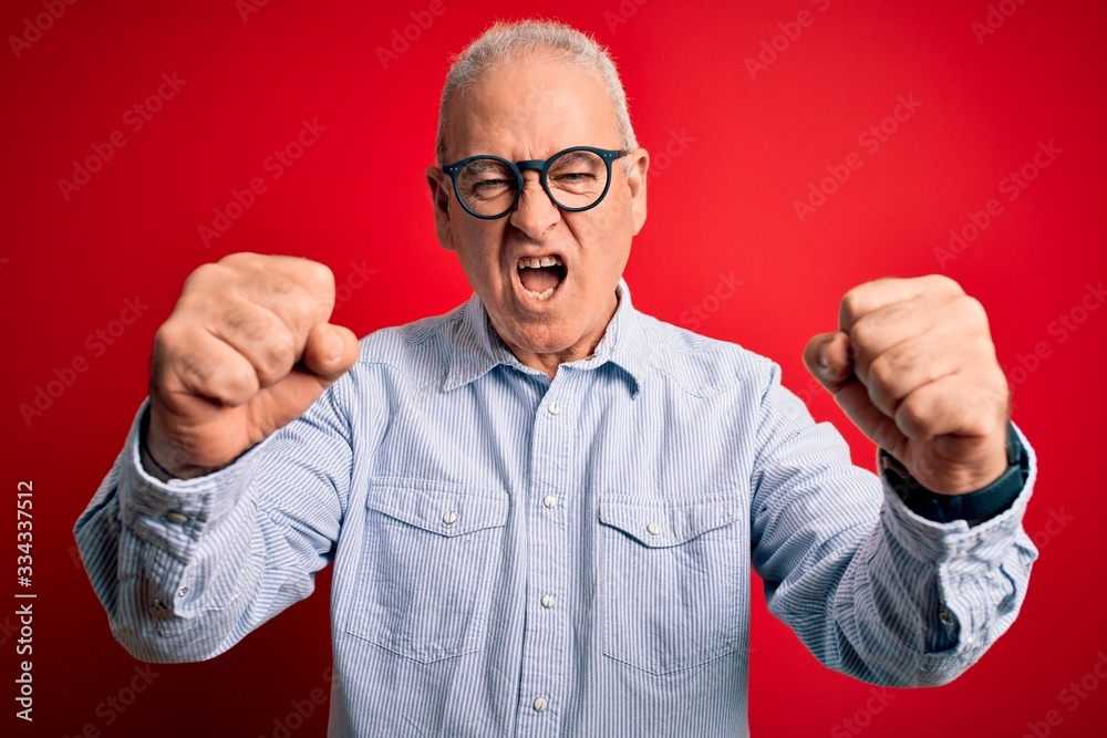 Middle age handsome hoary man wearing casual striped shirt and glasses over red background angry and mad raising fists frustrated and furious while shouting with anger. Rage and aggressive concept. Middle age handsome hoary man wearing casual striped shirt and glasses over red background angry and mad raising fists frustrated and furious while shouting with anger. Rage and aggressive concept.