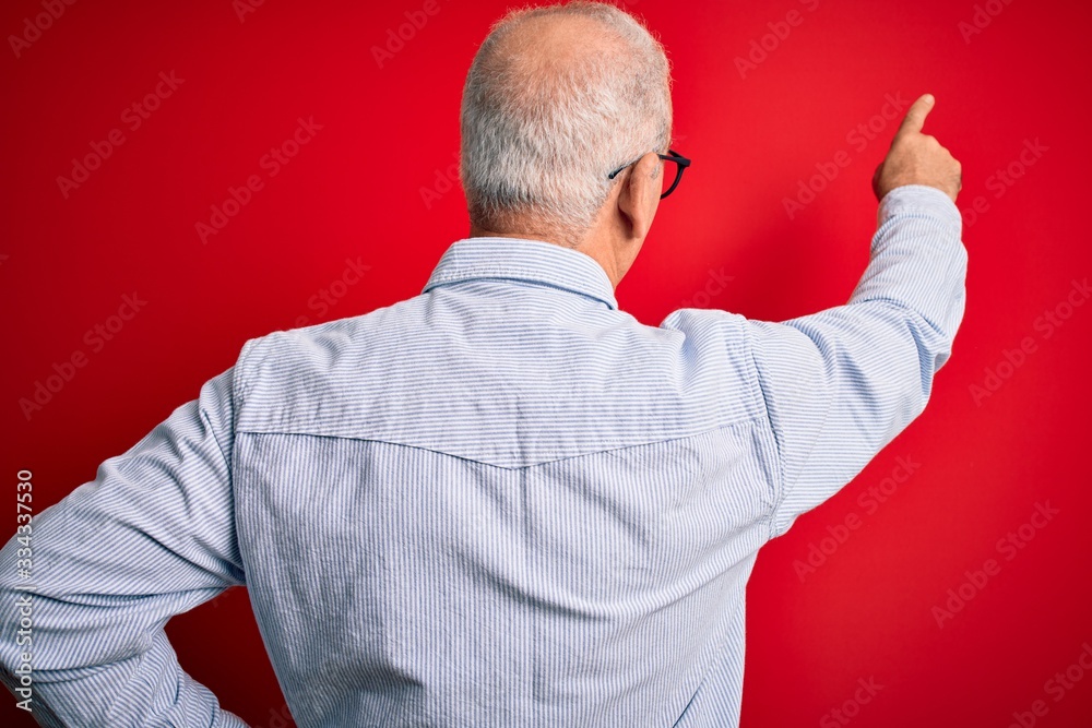 Middle age handsome hoary man wearing casual striped shirt and glasses over red background Posing backwards pointing ahead with finger hand Middle age handsome hoary man wearing casual striped shirt and glasses over red background Posing backwards pointing ahead with finger hand