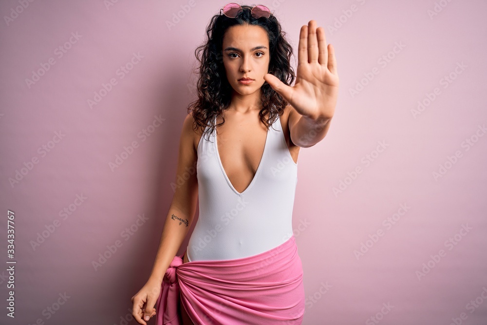 Beautiful woman with curly hair on vacation wearing white swimsuit over pink background doing stop sing with palm of the hand. Warning expression with negative and serious gesture on the face. Beautiful woman with curly hair on vacation wearing white swimsuit over pink background doing stop sing with palm of the hand. Warning expression with negative and serious gesture on the face.