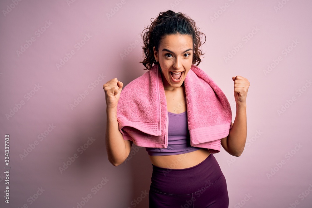 Young beautiful sportswoman with curly hair doing sport using towel over pink background celebrating surprised and amazed for success with arms raised and open eyes. Winner concept. Young beautiful sportswoman with curly hair doing sport using towel over pink background celebrating surprised and amazed for success with arms raised and open eyes. Winner concept.