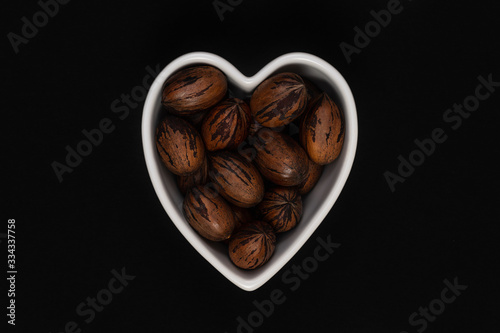 Pecan nuts with the nutshell in a heart shaped bowl on a black background. © VIS Fine Arts