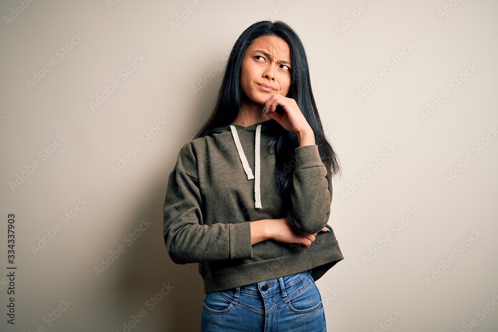 Young beautiful chinese sporty woman wearing sweatshirt over isolated white background with hand on chin thinking about question, pensive expression. Smiling with thoughtful face. Doubt concept. Young beautiful chinese sporty woman wearing sweatshirt over isolated white background with hand on chin thinking about question, pensive expression. Smiling with thoughtful face. Doubt concept.