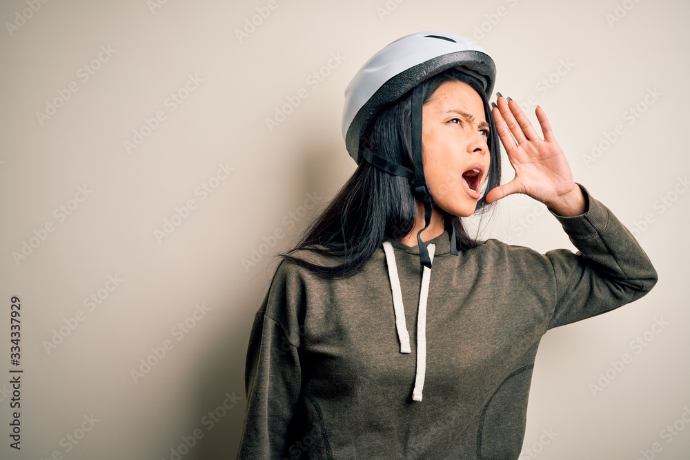 Young beautiful chinese woman wearing bike helmet over isolated white background shouting and screaming loud to side with hand on mouth. Communication concept. Young beautiful chinese woman wearing bike helmet over isolated white background shouting and screaming loud to side with hand on mouth. Communication concept.