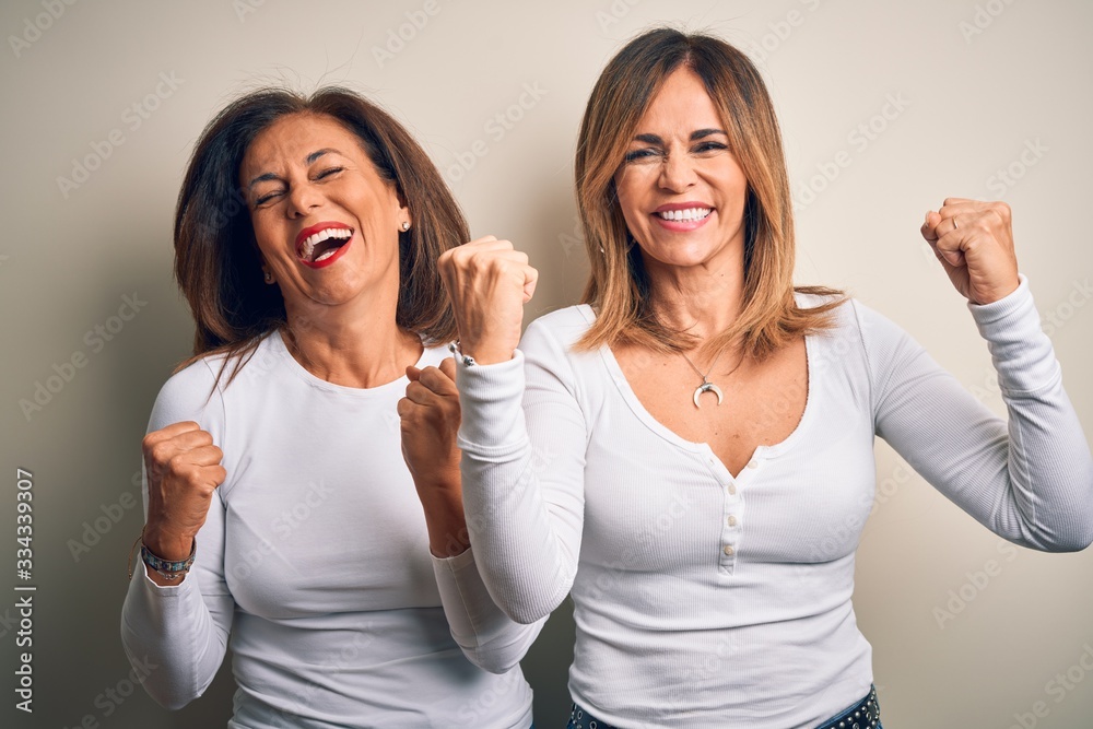 Middle age beautiful couple of sisters wearing casual t-shirt over isolated white background very happy and excited doing winner gesture with arms raised, smiling and screaming for success Middle age beautiful couple of sisters wearing casual t-shirt over isolated white background very happy and excited doing winner gesture with arms raised, smiling and screaming for success