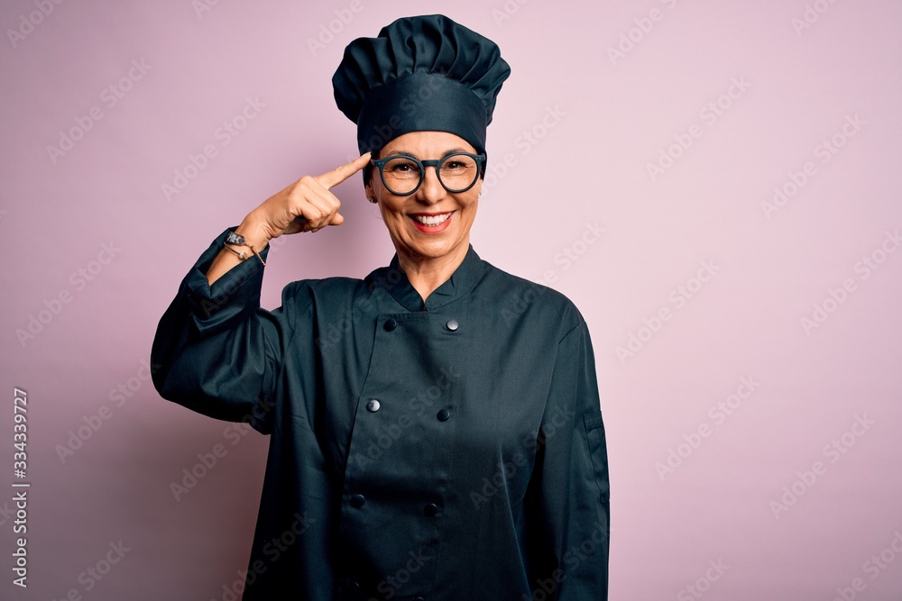 Middle age brunette chef woman wearing cooker uniform and hat over isolated pink background Smiling pointing to head with one finger, great idea or thought, good memory Middle age brunette chef woman wearing cooker uniform and hat over isolated pink background Smiling pointing to head with one finger, great idea or thought, good memory