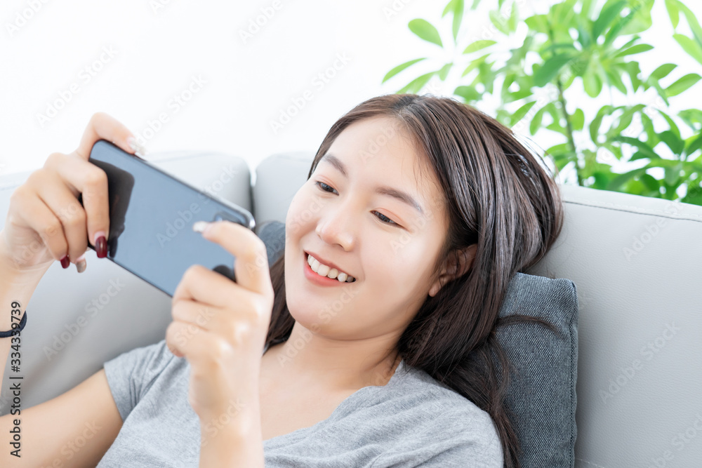 Attractive young Asian woman happy lie down on grey sofa in living room and using mobile phone connect internet for play game online Attractive young Asian woman happy lie down on grey sofa in living room and using mobile phone connect internet for play game online