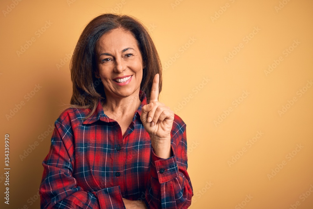 Middle age beautiful woman wearing casual shirt standing over isolated yellow background pointing finger up with successful idea. Exited and happy. Number one. Middle age beautiful woman wearing casual shirt standing over isolated yellow background pointing finger up with successful idea. Exited and happy. Number one.