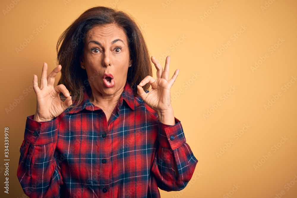 Middle age beautiful woman wearing casual shirt standing over isolated yellow background looking surprised and shocked doing ok approval symbol with fingers. Crazy expression Middle age beautiful woman wearing casual shirt standing over isolated yellow background looking surprised and shocked doing ok approval symbol with fingers. Crazy expression