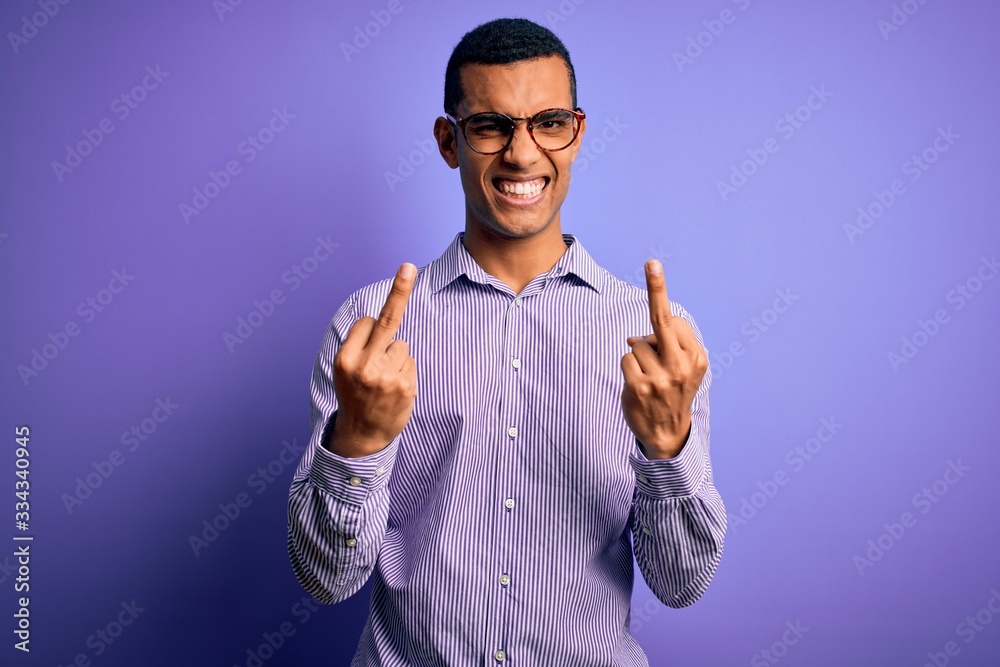 Fototapeta premium Handsome african american man wearing striped shirt and glasses over purple background Showing middle finger doing fuck you bad expression, provocation and rude attitude. Screaming excited