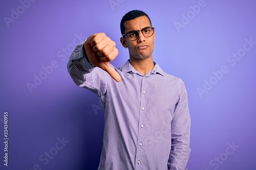 Handsome african american man wearing striped shirt and glasses over purple background looking unhappy and angry showing rejection and negative with thumbs down gesture. Bad expression.