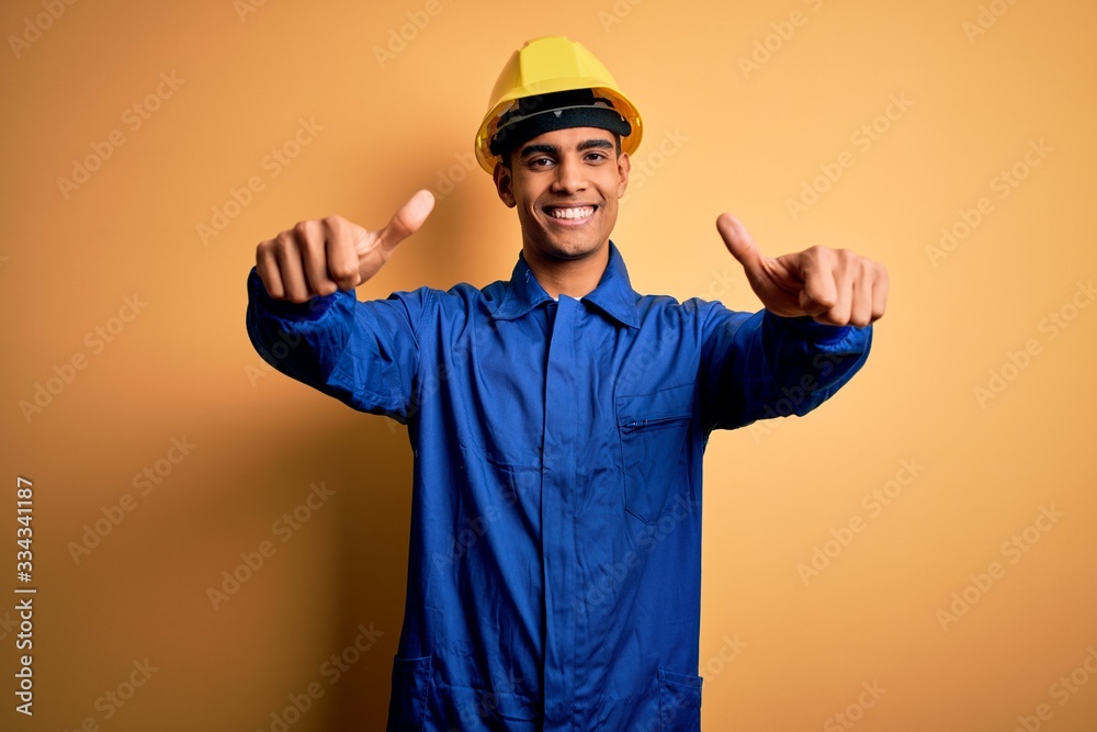 Young handsome african american worker man wearing blue uniform and security helmet approving doing positive gesture with hand, thumbs up smiling and happy for success. Winner gesture.