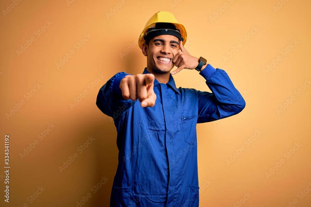 Young handsome african american worker man wearing blue uniform and security helmet smiling doing talking on the telephone gesture and pointing to you. Call me.