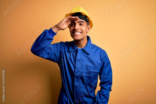 Young handsome african american worker man wearing blue uniform and security helmet very happy and smiling looking far away with hand over head. Searching concept.