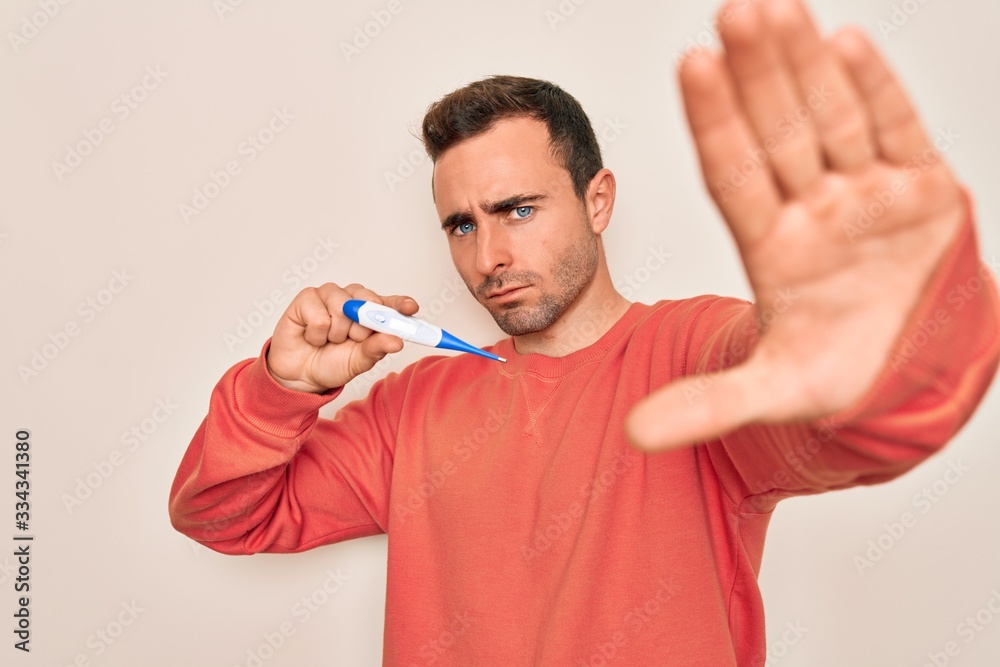 Young handsome man with blue eyes measuring temperature using digital thermometer with open hand doing stop sign with serious and confident expression, defense gesture Young handsome man with blue eyes measuring temperature using digital thermometer with open hand doing stop sign with serious and confident expression, defense gesture